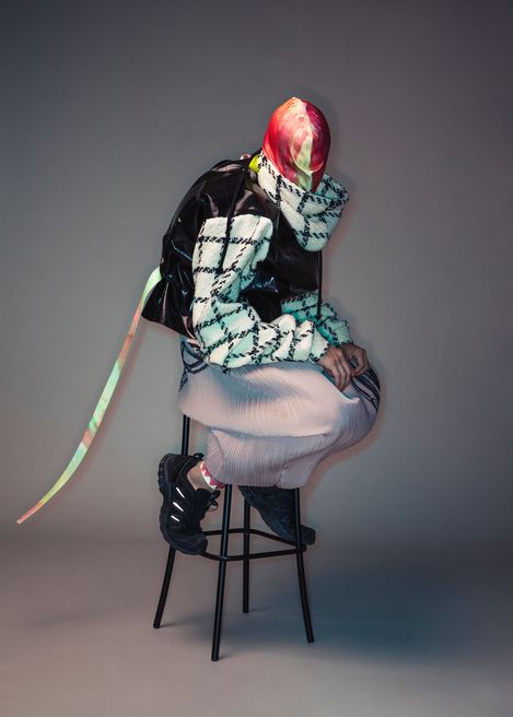 Person wearing a red-and-white patterned headscarf, checkered sweater, black vest, pleated skirt, and black shoes sitting on a stool leaning forward.