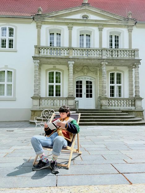 Person sitzt in legerer Kleidung auf einem Liegestuhl vor einem historischen Gebäude mit Säulen und Balkon.