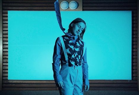 Person wearing a shirt, suspenders, and a tie standing in front of a light blue screen in a dark room.