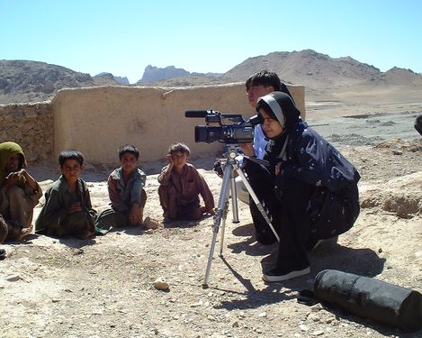 A person operates a video camera on a tripod in front of a group of children in a barren, mountainous landscape with a mud wall in the background.