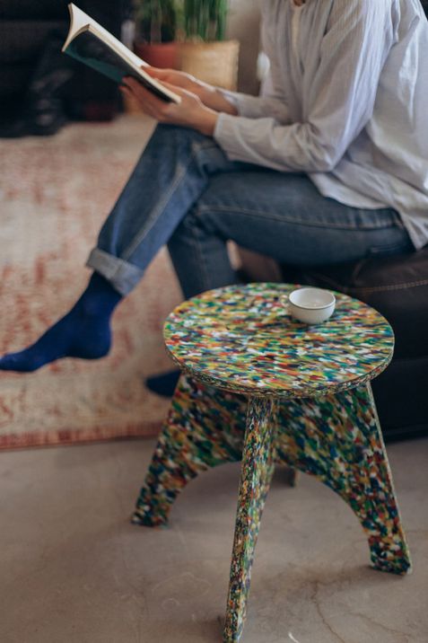 Person sitting on a sofa with crossed legs reading a book next to a small colorful stool with a white cup on it