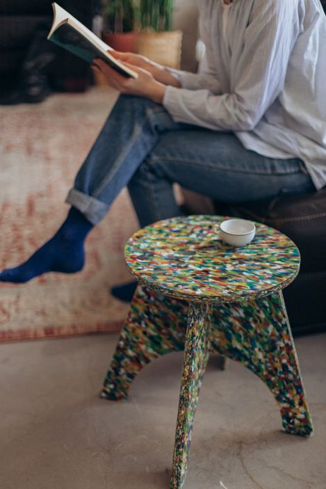 Person sitting on a sofa with crossed legs reading a book next to a small colorful stool with a white cup on it