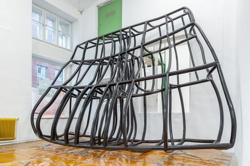 Sculpture made of black metal tubes in a room with windows and a green door forming a distorted bulging shape.