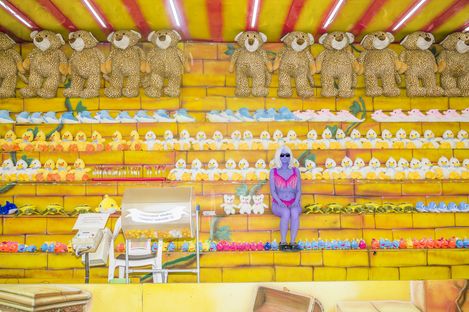Female figure in purple clothing and sunglasses sitting on yellow steps with rows of plush bears, ducks, and other stuffed animals.