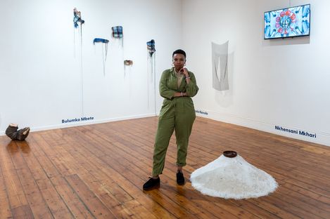 Person in green coveralls stands with arms crossed in a gallery with several wall installations and a white floor installation with a brown object.