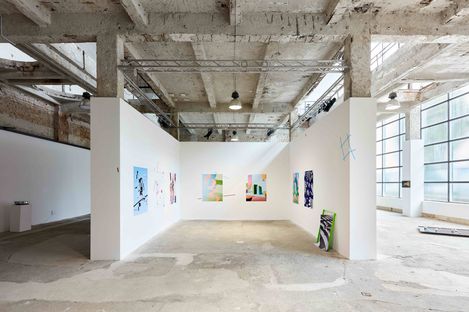 Large open exhibition space with raw concrete ceiling and white walls displaying several colorful abstract paintings.