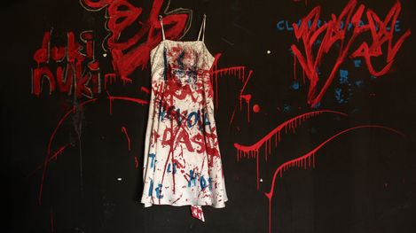 White dress splattered with red and blue paint hanging against a black wall with red and blue graffiti writing.