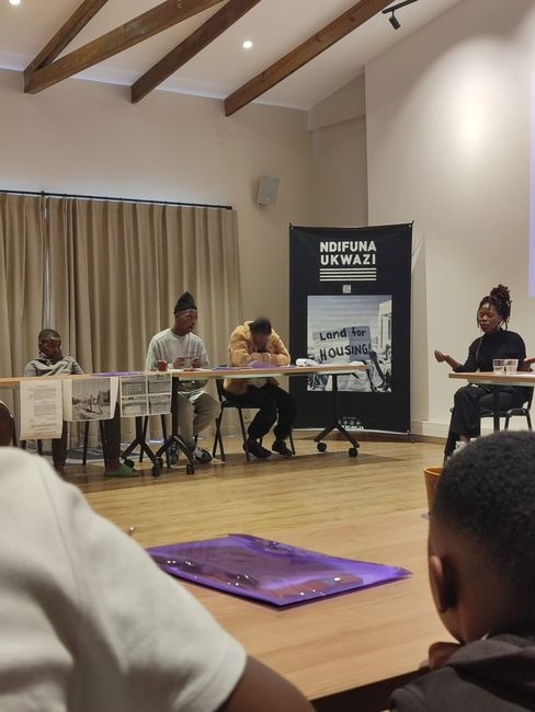 Four people are seated at two tables in a room with wooden floors and exposed ceiling beams. In the background, a black banner can be seen with the words "NDIFUNA UKWAZI" and "Land for Housing" written on it.