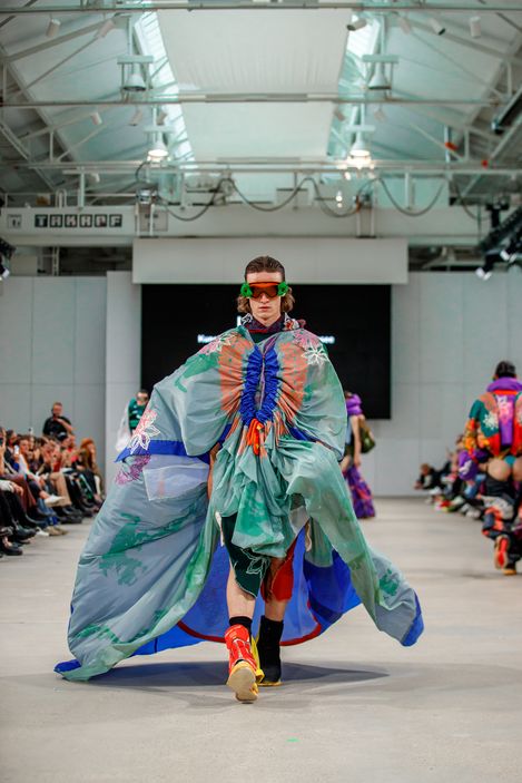 Person wearing a wide, colorful cape walking on a runway in a hall with audience on both sides.