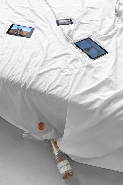 Three tablets lie on a wrinkled white bedsheet with a bottle of sparkling wine and a glass of rosé on the floor beneath.