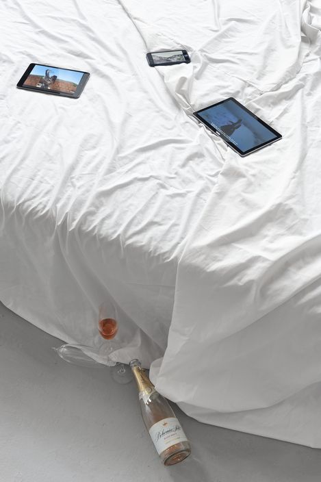 Three tablets lie on a wrinkled white bedsheet with a bottle of sparkling wine and a glass of rosé on the floor beneath.