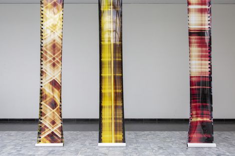 Three vertically hanging fabric panels with abstract plaid patterns in different color tones against a white wall and gray floor.