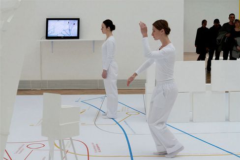 Two performers in white costumes perform movements within a minimalist exhibition installation, with visitors in the background.