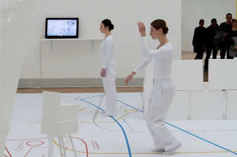 Two performers in white costumes perform movements within a minimalist exhibition installation, with visitors in the background.