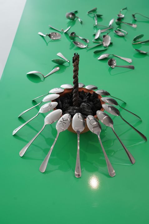 Anordnung von Metalllöffeln auf grünem Untergrund, die einen Kreis um einen in der Mitte stehenden, verkohlten Docht bilden, mit weiteren verstreuten Löffeln im Hintergrund.
