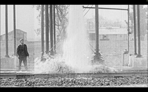 Mann steht neben einer großen Wasserfontäne in einem offenen, von Säulen getragenen Bauwerk.