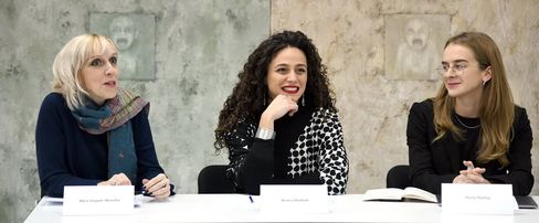 Drei Frauen sitzen an einem Tisch mit Namensschildern vor einer hellen Wand mit zwei Reliefs.