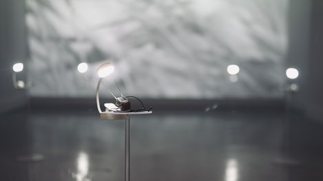 A small object with a round light on a thin stand against a blurred bright background.