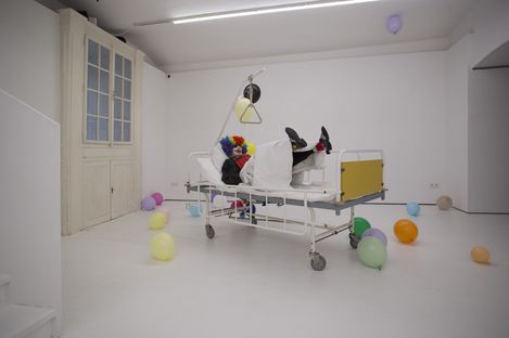 Person wearing a colorful clown wig lying on a hospital bed in a white room with scattered balloons.