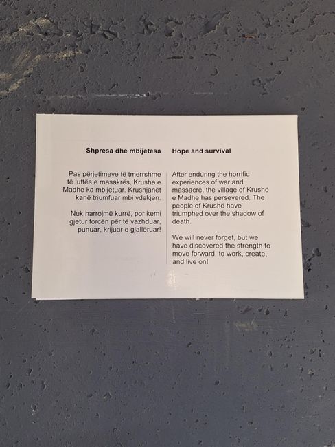 White sign with bilingual text in Albanian and English about hope and survival after war and massacre.