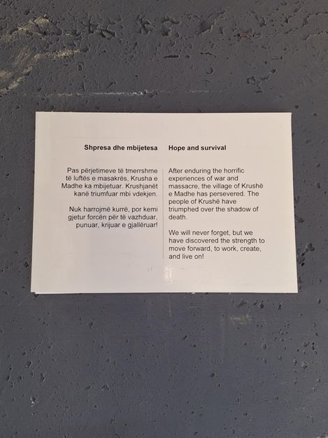 White sign with bilingual text in Albanian and English about hope and survival after war and massacre.