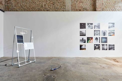 Art gallery with a white wall displaying a grid of photographs and a freestanding metal structure with a screen and a white sign on a concrete floor.