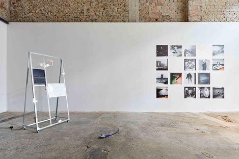 Art gallery with a white wall displaying a grid of photographs and a freestanding metal structure with a screen and a white sign on a concrete floor.