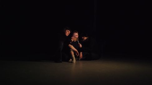 Three people sitting close together on the floor in a dark room, one person holding a smartphone.