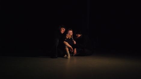 Three people sitting close together on the floor in a dark room, one person holding a smartphone.