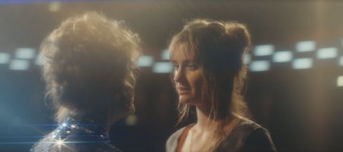 Zwei Personen mit hochgesteckten Haaren stehen sich in unscharfer Aufnahme gegenüber vor einem Hintergrund mit hellen, rechteckigen Lichtquellen.