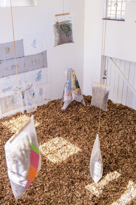 Room with floor covered in wood chips and several hanging transparent bags filled with various materials and a painted cloth on a stand.