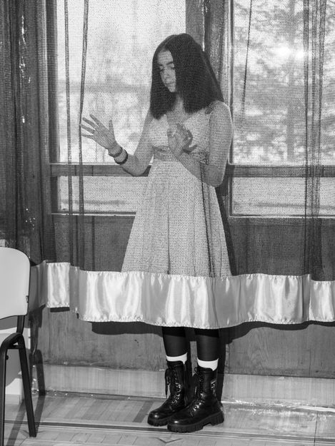 Woman with long hair and dress standing behind a semi-transparent curtain gesturing with her hands.