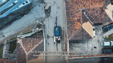 Aerial view of a residential area with red tiled roofs, a street, and several parked cars including a blue truck.