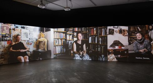 Three projected portraits of seated people in a dark room with visible books, dolls, and other objects in the background.