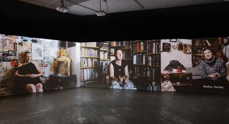 Three projected portraits of seated people in a dark room with visible books, dolls, and other objects in the background.
