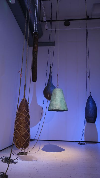Several differently shaped and sized objects hanging from the ceiling by ropes in a room with white walls and wooden floor.