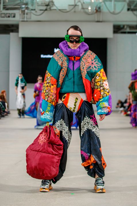 Person on runway wearing colorful padded jacket with floral patterns, wide black pants with lace appliqués, holding large red bag and wearing green headphones.