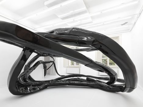 Black glossy sculpture with curved, intertwined organic shapes in a white room with windows.