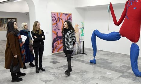 Vier Frauen stehen in einer Galerie vor einer großen, abstrakten Skulptur mit roten und blauen Elementen und hören einer Frau mit schwarz-weiß kariertem Mantel zu.