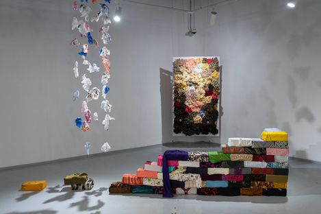 Installation with hanging plastic fragments and stacked fabric-covered objects in a gallery space.
