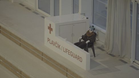 Person wearing a panda mask and black clothing sitting on a chair behind a sign reading "PLAVČÍK | LIFEGUARD" at a swimming pool.