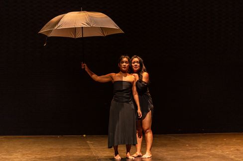 Zwei Frauen in schwarzen Kleidern stehen barfuß auf einer Bühne, eine hält einen geöffneten Regenschirm über beide.