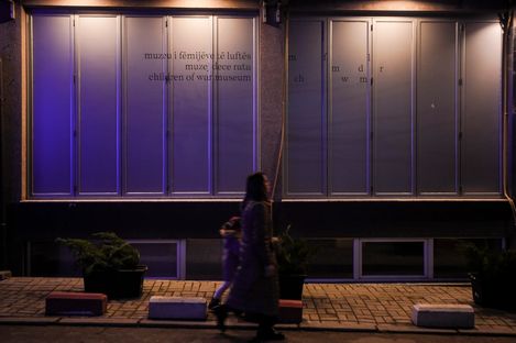 A woman and a child walk past a building at night with large closed windows and partially legible text.