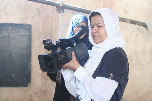 Two women, one holding a video camera, the other standing behind her in a room with a chalkboard on the wall.