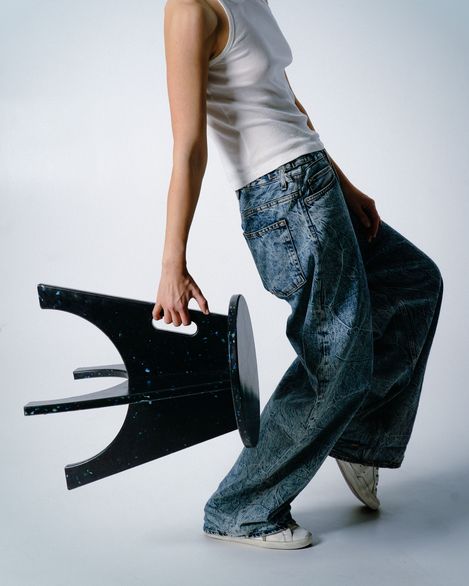 Person wearing loose jeans and a white tank top holding a black chair tilted sideways downward