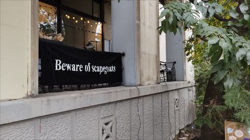 Black banner with white text "Beware of scapegoats" attached to a building facade with windows and plants in front.