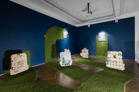 Exhibition room with dark blue walls, green carpet strips on the floor and wall, and several sculptural objects featuring floral and geometric patterns.