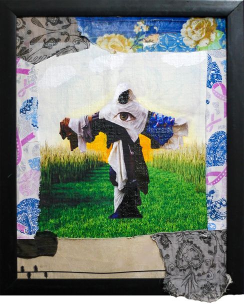 The image shows a framed mixed-media textile artwork featuring a central surreal figure. The figure stands on a green field, has outstretched arms, and an eye in the head area. Various fabrics, patterns, and collage elements form the background and border. The piece has a dreamlike, symbolic quality.