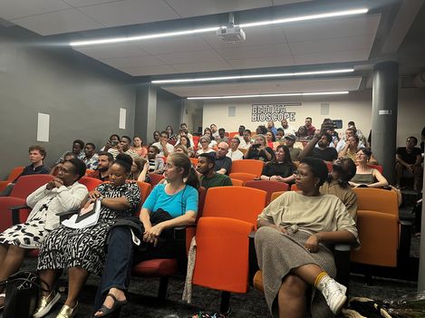 Several people sitting in a small cinema with orange and red seats, a sign reading "BERLINA BIOSCOPE" visible in the background.