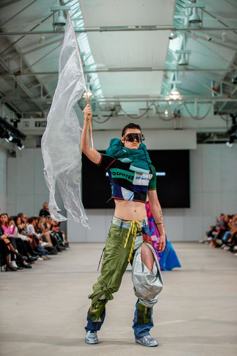 Person on runway wearing green pants with silver patch, cropped top, and holding a large white flag.